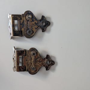 May include: Two antique silver-toned hinges with ornate, decorative designs. Each hinge has a rectangular section with multiple screw holes and a stylized, curved end with circular cutouts. The hinges are likely for a cabinet or small box.