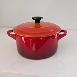 May include: A round enameled cast iron Dutch oven with a gradient finish transitioning from a deep cherry red at the base to a bright orange on the lid. It features two side handles and a black circular knob centered on the lid.