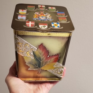 May include: A vintage metal tin with a rectangular shape and a hinged lid. The tin features a large, colorful maple leaf design on the front and various crests on the lid. The tin has a gold and brown color scheme.