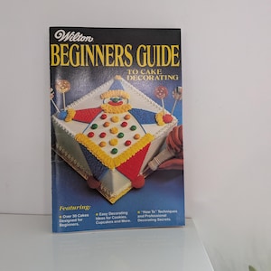May include: A Wilton Beginners Guide to Cake Decorating softcover book with a black cover featuring yellow text and a photograph of a square cake decorated like a clown with red, blue, and yellow frosting, colorful candies, and lollipops.
