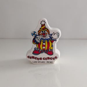 Puede incluir: Figura de cerámica blanca de un payaso sosteniendo un caramelo. El payaso tiene una nariz roja, zapatos rojos y un atuendo colorido. El texto "Circus Circus" y "Las Vegas • Reno" está impreso en la base.