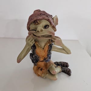 Könnte beinhalten: Eine hellgrüne Koboldfigur mit einer dunkelroten Strickmütze, einer dunkelblauen Weste und einer orangefarbenen Tunika. Der Kobold sitzt und zieht mit beiden Händen die Mundwinkel weit auseinander, was einen verspielten Gesichtsausdruck erzeugt.