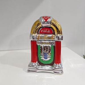 May include: A ceramic jukebox figurine featuring a shiny silver finish with red and green accents. The top displays the Coca-Cola logo in white text on a red background. The centre section includes the text Rock-n-Roll in gold script lettering.