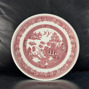 Peut inclure: Une assiette ronde blanche avec un motif Willow rouge. L'assiette présente une scène de paysage détaillée avec des pagodes, des ponts et des oiseaux. Le bord a une bordure décorative. L'assiette est posée sur un fond sombre.