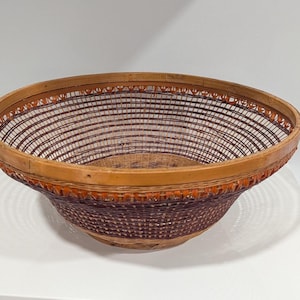 Puede incluir: Una cesta tejida a mano con un diseño ancho y poco profundo. La cesta presenta un borde de madera natural y un cuerpo tejido con hebras marrones oscuras y claras alternas. El borde tiene detalles decorativos en naranja. La cesta se utiliza probablemente para guardar o exhibir.