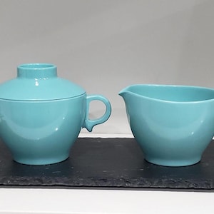 May include: A turquoise ceramic sugar bowl with a lid and a creamer with a handle. The matching set is displayed on a dark gray rectangular tray. The sugar bowl has two handles and a lid with a small knob.