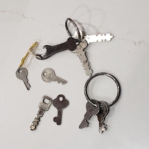 May include: A collection of vintage keys, including a bottle opener key, arranged on a white surface. The keys are various sizes and shapes, with a mix of silver and dark metal finishes. Some keys are attached to metal rings.