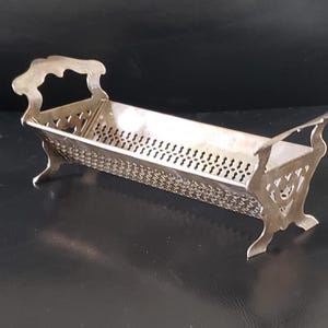 Puede incluir: Una bandeja rectangular de color plateado con un diseño calado decorativo. La bandeja tiene un asa en cada extremo y descansa sobre cuatro patas ornamentadas. La bandeja mide aproximadamente 25 cm de largo.