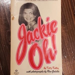 Könnte beinhalten: Ein Taschenbuch mit dem Titel "Jackie Oh!" mit einem Foto einer lächelnden Frau. Der Titel ist in großen rosa Buchstaben geschrieben. Das Cover enthält auch Text darüber, dass das Buch ein Bestseller ist und den Autor.