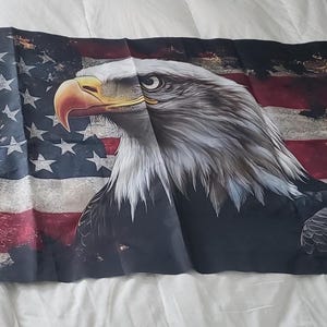 Könnte beinhalten: Eine Flagge mit der amerikanischen Flagge und einem detaillierten Bild eines Weißkopfseeadlers. Der Adler hat weiße und graue Federn, einen gelben Schnabel und ist vor dem Hintergrund der rot-weiß-blauen Flagge abgebildet.