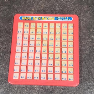 May include: A red plastic Magic Math Machine for addition and subtraction. The board has a grid of white tiles with math problems like 1+1, 2-1, and 3+3. The text "MAGIC MATH MACHINE ADDITION & SUBTRACTION" is at the top.