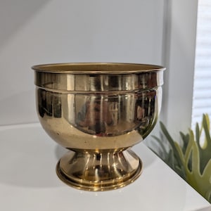 May include: A polished brass bowl with a smooth, rounded body and a wide, flared rim. The bowl features a pedestal base with a circular foot, creating a stable foundation. The metallic surface is reflective, showing a warm, golden tone throughout.