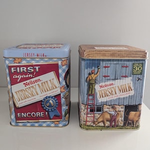 May include: Two rectangular vintage Jersey Milk chocolate tins. One tin has a red and blue design with the text "First again!" and "Encore!". The other tin shows a barn scene with a worker painting and cows.