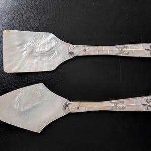 Könnte beinhalten: Zwei Servierutensilien: ein Pfannenwender und ein Tortenheber. Beide haben Perlmuttklingen und Griffe mit silbernen Akzenten und dekorativen Endstücken. Der Pfannenwender ist rechteckig, und der Tortenheber hat eine dreieckige Klinge.