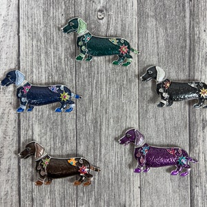 May include: Five dachshund dog pins with colourful enamel designs. The pins are black, green, blue, purple, and brown with white accents. Each pin has a different floral design.