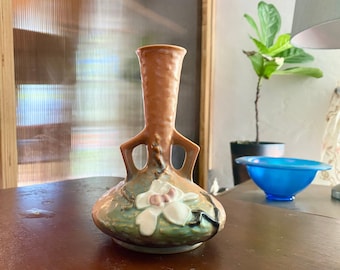 May include: A brown and green ceramic vase with a white flower design. The vase has a unique shape with two handles and a wide base.