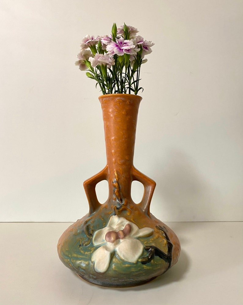 Może przedstawiać: Brązowy i zielony, glazurowany ceramiczny wazon z dwoma uchwytami i kwiatowym wzorem. Wazon jest ozdobiony białym kwiatem magnolii z r&oacute;żowymi akcentami. Wazon jest wypełniony r&oacute;żowymi i białymi goździkami.