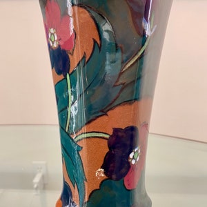 K&ouml;nnte beinhalten: Eine gr&uuml;ne und braune Keramikvase mit geometrischer Form und Blumenmuster. Die Vase zeigt rote, gr&uuml;ne, blaue und wei&szlig;e Blumen.