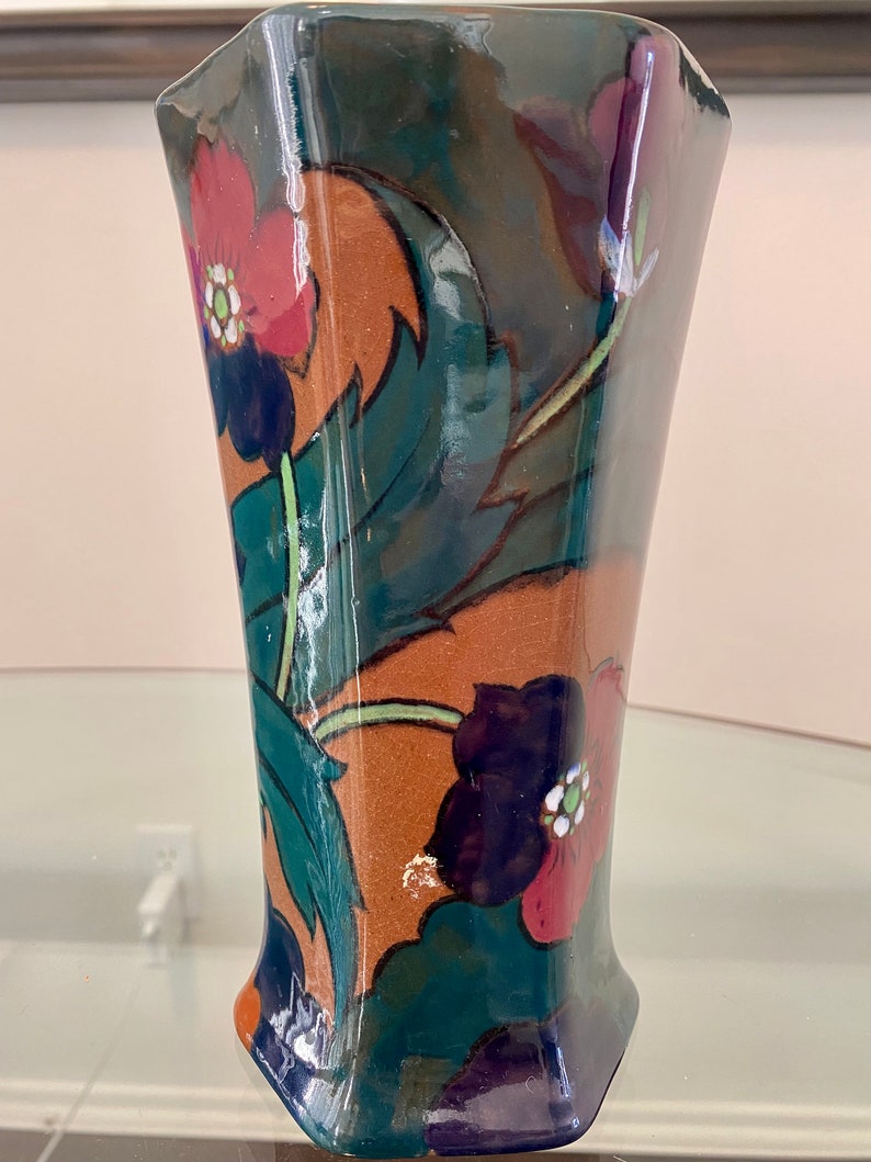 K&ouml;nnte beinhalten: Eine gr&uuml;ne und braune Keramikvase mit geometrischer Form und Blumenmuster. Die Vase zeigt rote, gr&uuml;ne, blaue und wei&szlig;e Blumen.
