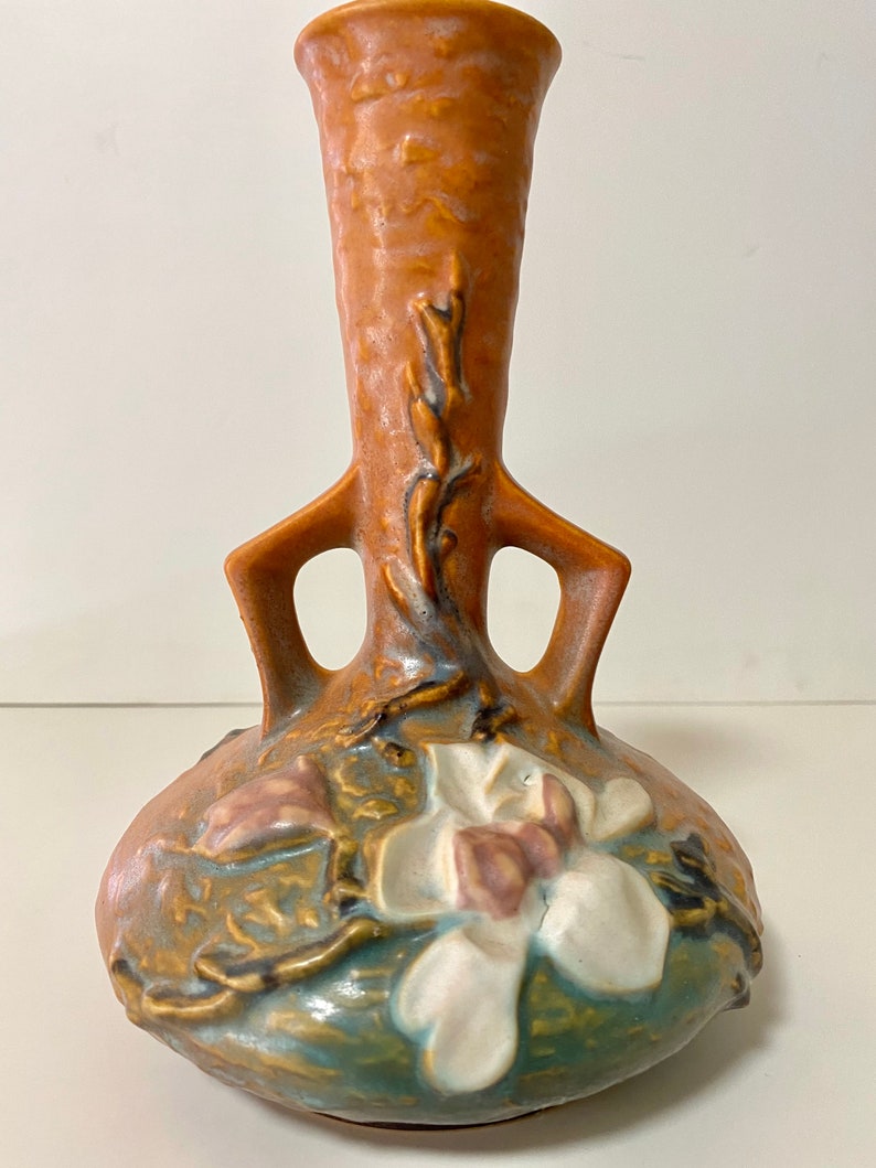 Może przedstawiać: Brązowy i niebieski ceramiczny wazon z wzorem białego kwiatu magnolii i dwoma uchwytami. Wazon ma fakturowaną powierzchnię i wąską szyjkę.