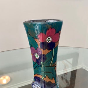 K&ouml;nnte beinhalten: Eine kleine, gr&uuml;ne, blaue und braune Keramikvase mit geometrischer Form und floralem Design. Die Vase zeigt rote, violette und wei&szlig;e Blumen mit gr&uuml;nen Bl&auml;ttern.
