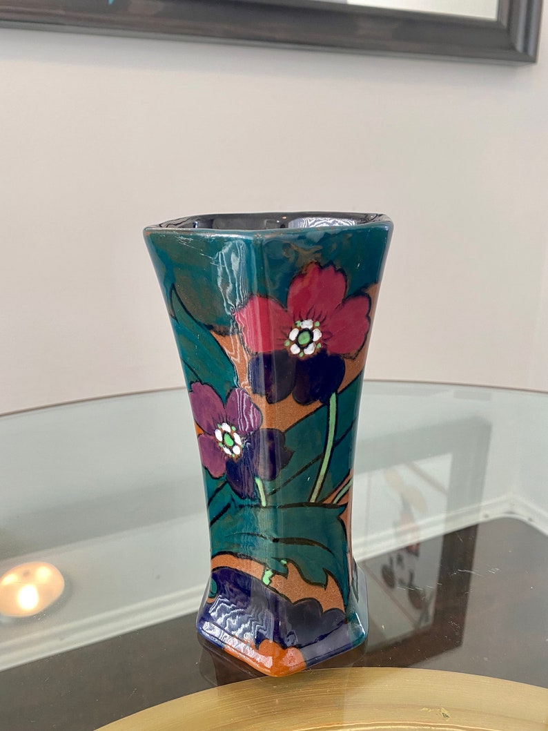 K&ouml;nnte beinhalten: Eine kleine, gr&uuml;ne, blaue und braune Keramikvase mit geometrischer Form und floralem Design. Die Vase zeigt rote, violette und wei&szlig;e Blumen mit gr&uuml;nen Bl&auml;ttern.
