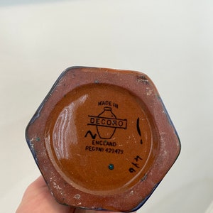 K&ouml;nnte beinhalten: Eine braune sechseckige Keramikvase mit blauer Glasur. Der Boden der Vase hat ein braunes Etikett mit dem Text "MADE IN DECORO ENGLAND REG.NO. 429479" und eine handgeschriebene Nummer "9143".