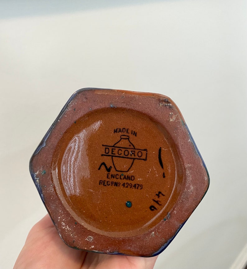 K&ouml;nnte beinhalten: Eine braune sechseckige Keramikvase mit blauer Glasur. Der Boden der Vase hat ein braunes Etikett mit dem Text "MADE IN DECORO ENGLAND REG.NO. 429479" und eine handgeschriebene Nummer "9143".