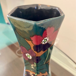 K&ouml;nnte beinhalten: Eine dunkelgr&uuml;ne Keramikvase mit geometrischer Form und floralem Design. Die Vase zeigt rote, violette und wei&szlig;e Blumen mit gr&uuml;nen Bl&auml;ttern.
