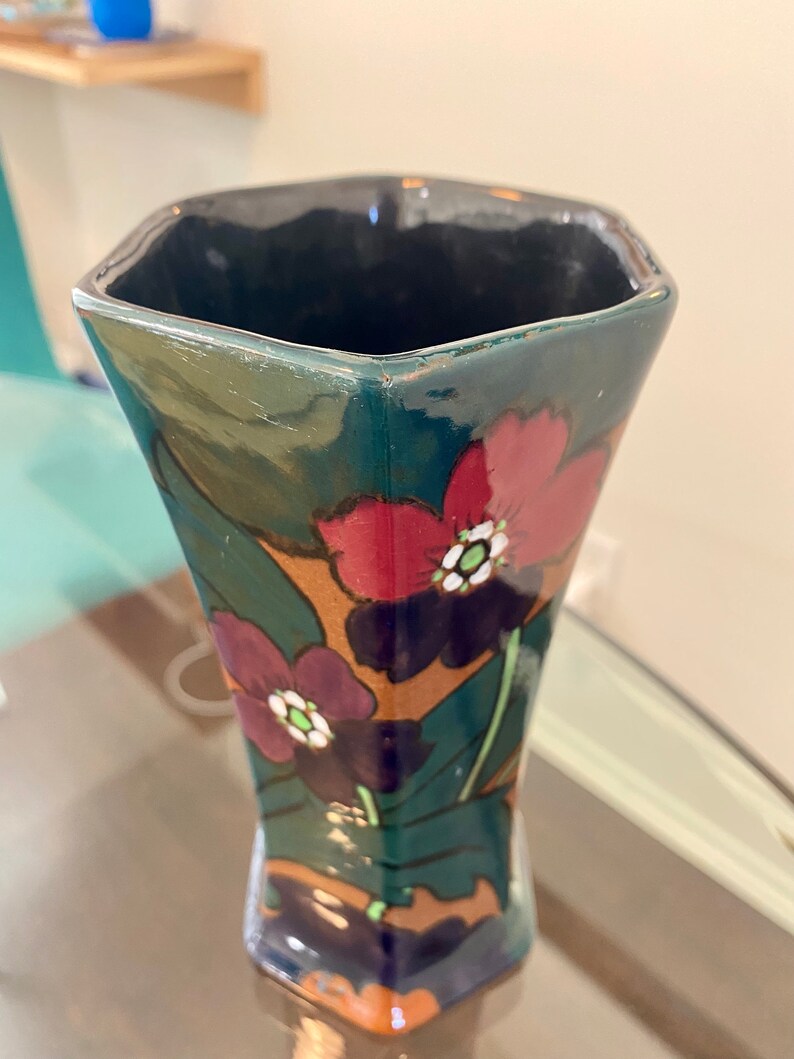 K&ouml;nnte beinhalten: Eine dunkelgr&uuml;ne Keramikvase mit geometrischer Form und floralem Design. Die Vase zeigt rote, violette und wei&szlig;e Blumen mit gr&uuml;nen Bl&auml;ttern.