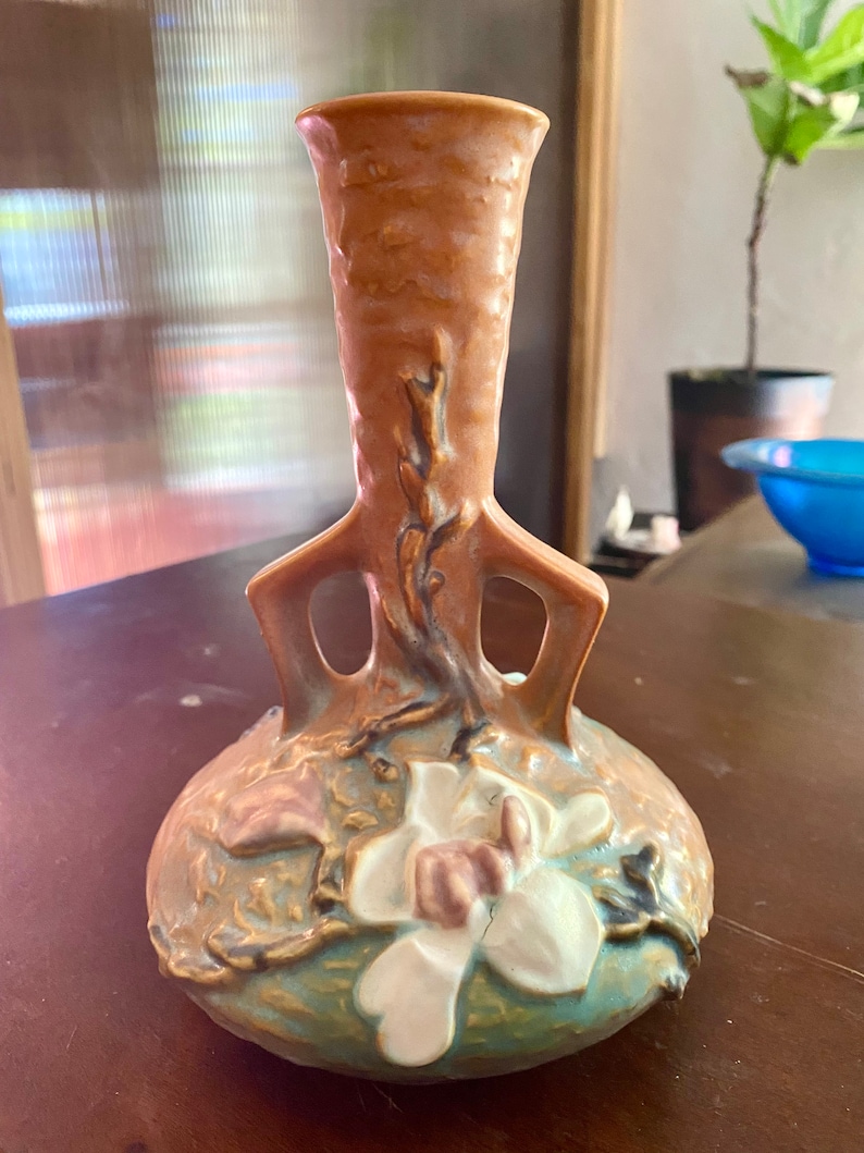 Może przedstawiać: Brązowy i zielony ceramiczny wazon z wzorem białego kwiatu magnolii. Wazon ma dwa uchwyty i wąską szyjkę.