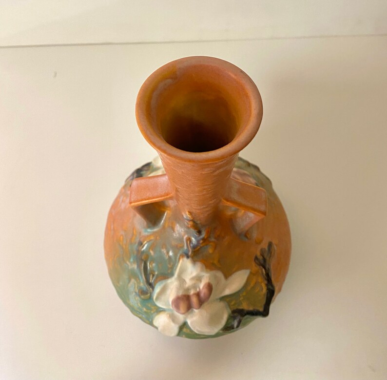 Może przedstawiać: Brązowy ceramiczny wazon z szeroką podstawą i wąską szyjką. Wazon jest ozdobiony białym kwiatem magnolii i zielonymi liśćmi.