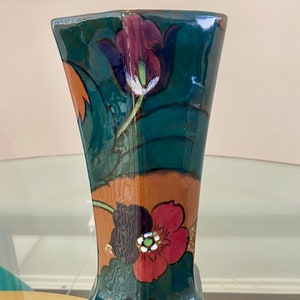 K&ouml;nnte beinhalten: Eine gr&uuml;ne und braune Keramikvase mit Blumenmuster. Die Vase hat eine geometrische Form und ist mit roten, wei&szlig;en und violetten Blumen verziert.