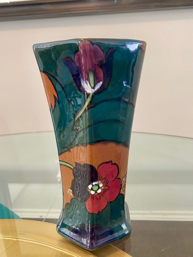 K&ouml;nnte beinhalten: Eine gr&uuml;ne und braune Keramikvase mit Blumenmuster. Die Vase hat eine geometrische Form und ist mit roten, wei&szlig;en und violetten Blumen verziert.