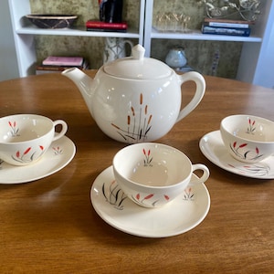 May include: A white teapot and four teacups with a floral design of red and brown reeds on a white background. The teapot has a lid and a handle. The teacups have handles and saucers.