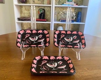 Op de afbeelding: Drie zwarte dienbladen met roze olifanten en een cocktailglasontwerp. De dienbladen staan op heldere acrylstandaards.