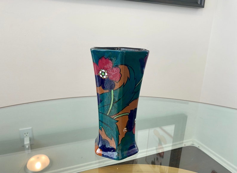K&ouml;nnte beinhalten: Eine tealblaue Keramikvase mit einem Blumenmuster. Die Vase hat eine geometrische Form und zeigt rote, blaue, gr&uuml;ne und orangefarbene Blumen.