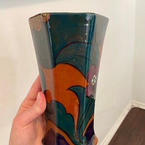K&ouml;nnte beinhalten: Eine Keramikvase mit geometrischer Form und einem farbenfrohen Blumendesign. Die Vase ist in Gr&uuml;n-, Blau-, Orange- und Violettt&ouml;nen bemalt.