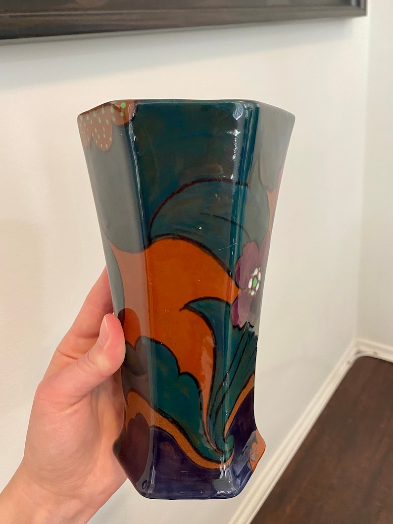 K&ouml;nnte beinhalten: Eine Keramikvase mit geometrischer Form und einem farbenfrohen Blumendesign. Die Vase ist in Gr&uuml;n-, Blau-, Orange- und Violettt&ouml;nen bemalt.