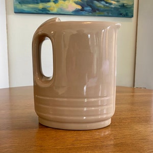 May include: A beige ceramic pitcher with a handle and a smooth, glossy finish. The pitcher has a rounded body and a wide mouth.
