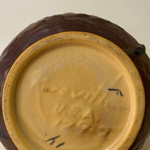Może przedstawiać: Sp&oacute;d brązowej, szkliwionej ceramicznej wazony z jasnobrązową krawędzią. Wazon ma znak producenta, kt&oacute;ry brzmi "Rookwood" i numer "1".