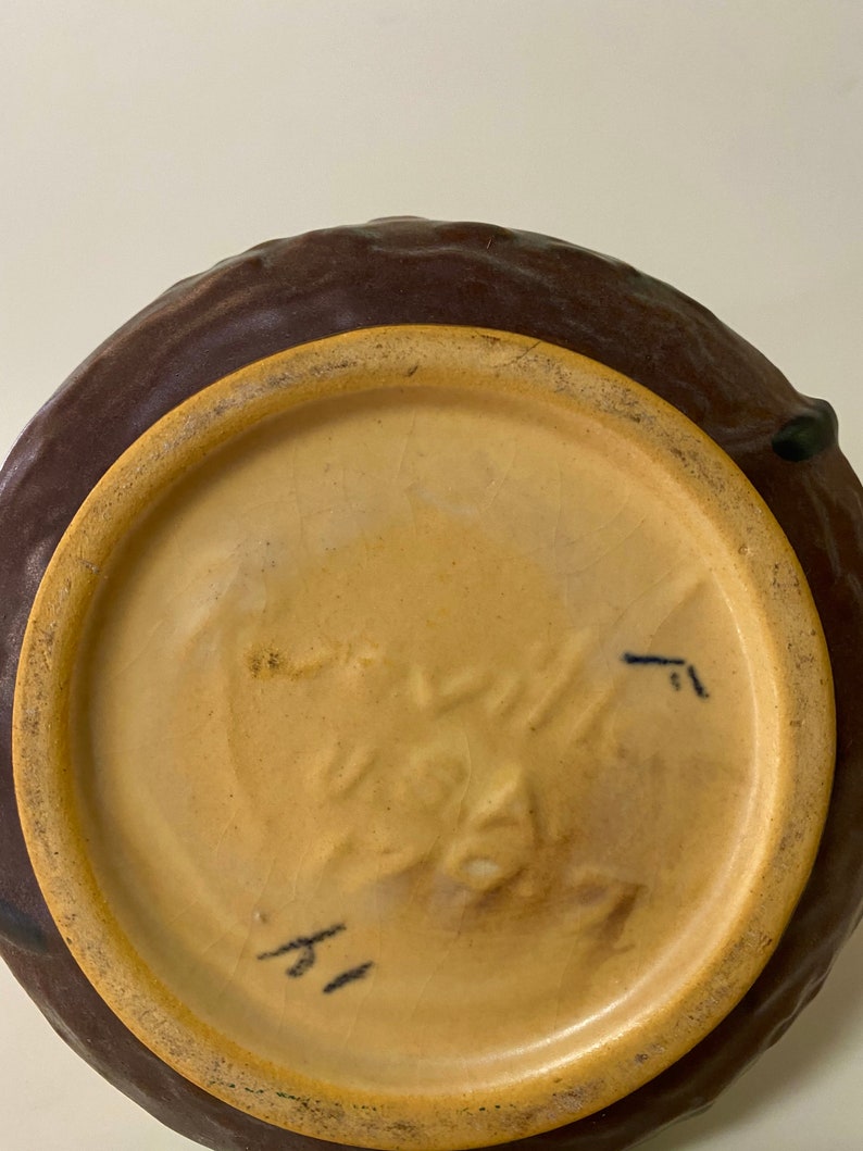 Może przedstawiać: Sp&oacute;d brązowej, szkliwionej ceramicznej wazony z jasnobrązową krawędzią. Wazon ma znak producenta, kt&oacute;ry brzmi "Rookwood" i numer "1".