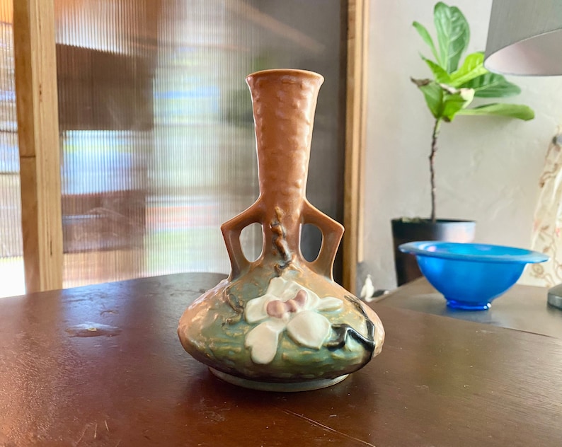 Może przedstawiać: Brązowy i zielony ceramiczny wazon z białym wzorem kwiatowym. Wazon ma unikalny kształt z dwoma uchwytami i szeroką podstawą.