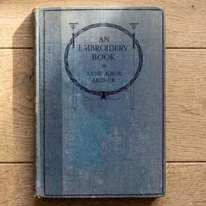 May include: A vintage hardcover book titled An Embroidery Book by Anne Knox Arthur. The cover is a faded blue fabric with a dark blue circular decorative border and black text centered on the front. The book has a rectangular shape and spine.