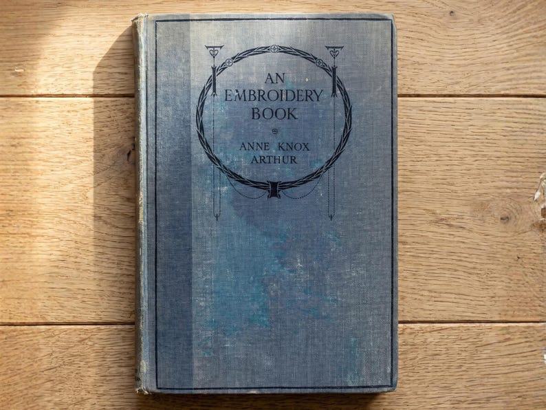 May include: A vintage hardcover book titled An Embroidery Book by Anne Knox Arthur. The cover is a faded blue fabric with a dark blue circular decorative border and black text centered on the front. The book has a rectangular shape and spine.
