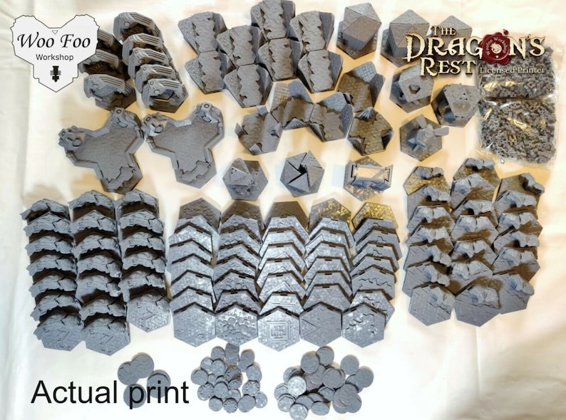 May include: A collection of gray 3D printed terrain pieces for tabletop gaming. The set includes various shapes and sizes of walls, towers, and other structures. The pieces are arranged on a white surface. The text "The Dragon's Rest" is visible in the upper right corner of the image. The text "Woo Foo Workshop" is visible in the upper left corner of the image.