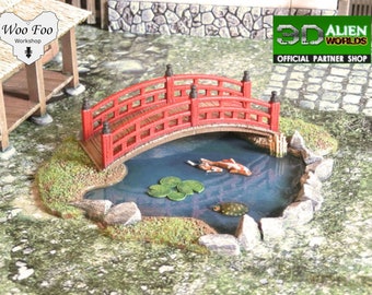 May include: A red wooden bridge arches over a small pond with a green lily pad and a turtle. Two orange and white fish swim in the pond. The pond is surrounded by rocks and green grass.