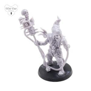 Puede incluir: Una figura en miniatura de plástico gris de una criatura parecida a un goblin que sostiene un micrófono y un bastón con un cráneo en la parte superior. La figura está de pie sobre una base negra.