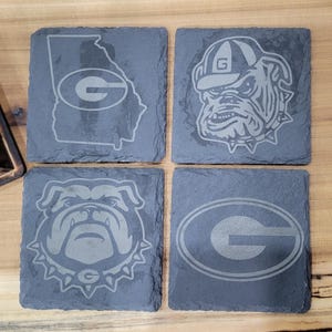 May include: Four black slate coasters with a white outline of the state of Georgia, a bulldog mascot, and the letter "G" in a circle.