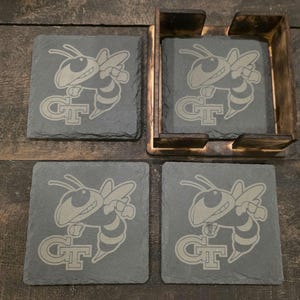 May include: Set of four square slate coasters with a laser-etched design of a yellow jacket and the letters "GT". The coasters are displayed with a wooden holder.